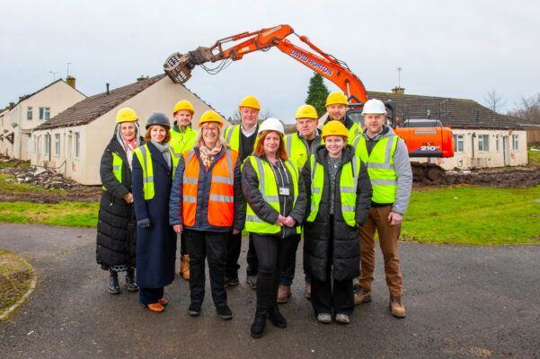 Group of people standing in front of houses as a digger pulls the houses down.