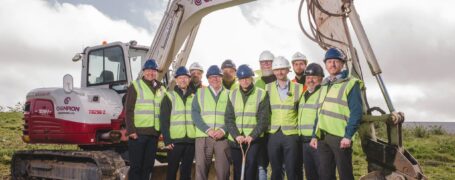 People in hi viz and hard hats under the arm of a digger