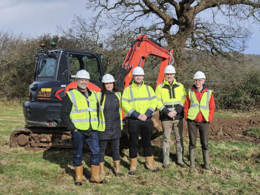 5 people standing a field in front of a digger