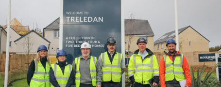 6 People wearing hard hats and hi viz standing in front of a sign for a new development