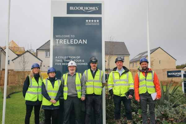 6 People wearing hard hats and hi viz standing in front of a sign for a new development